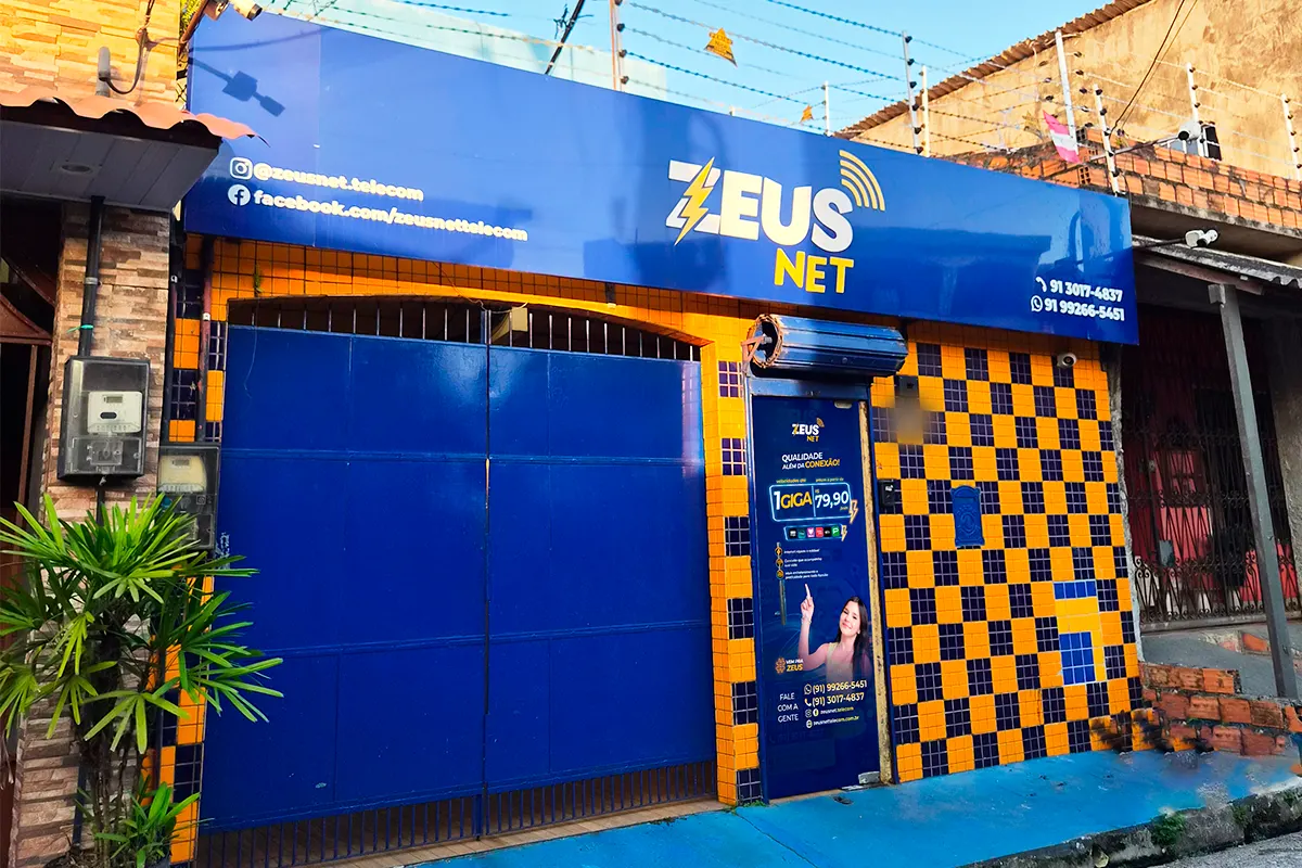 Fachada da loja da Zeus Net: letreiro azul com a marca, portão azul e parede de ladrilho amarelo e azul.
